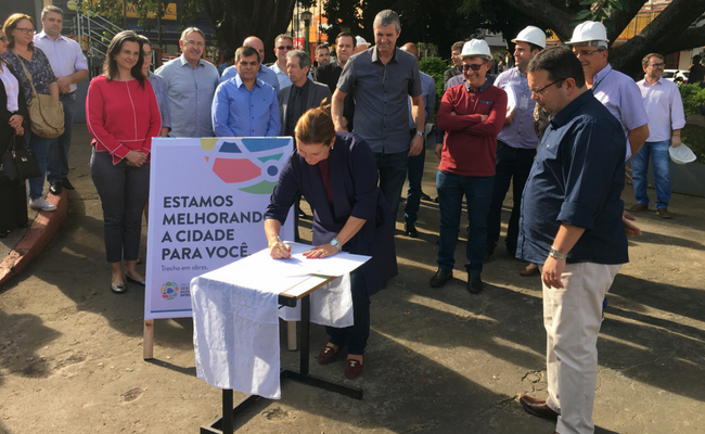 Prefeita Fátima em pé assina o documento que está em uma mesa. Público ao redor assiste a assinatura 