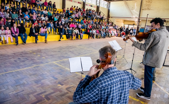 À direita, um músico em pé toca um violino e outro sentado toca um violoncelo. À esquerda, os alunos sentados em uma arquibancada