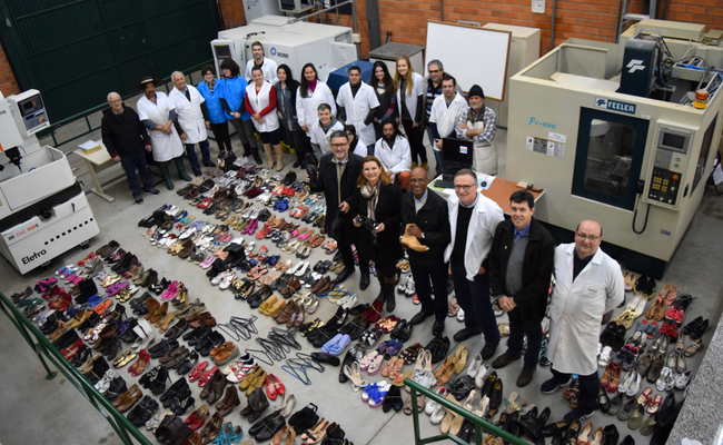 De uma ângulo de cima para baixo, sapatos estão no chão e a prefeita em pé no meio dos sapatos, juntamente com o restante da equipe 