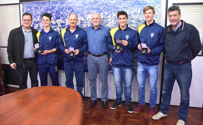 Sete homens em pé, lado a lado, com os três atletas e o técnico com as medalhas nas mãos