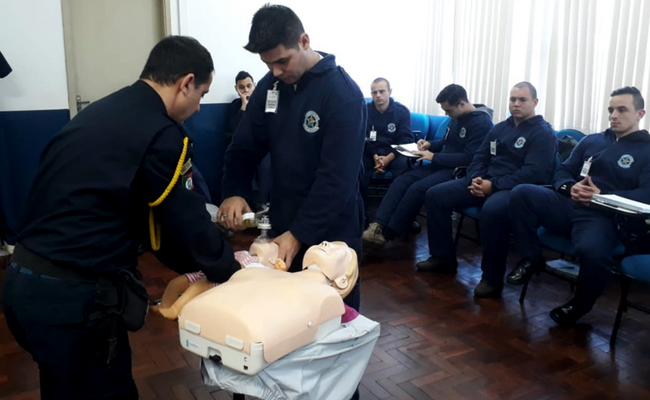Em pé, instrutor mostra a um aluno, que está em pé a sua frente, os movimentos corretos dos primeiros socorros em uma boneco, que está em cima de uma mesa. Ao lado, sentados, outros guardas acompanham o treinamento