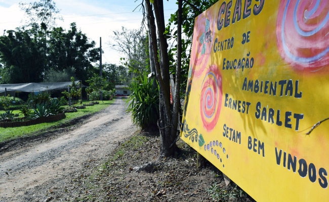À esquerda, uma rua de chão batido. À direita, uma placa com fundo amarelo escrito o Centro Municipal de Educação Ambiental Ernest Sarlet