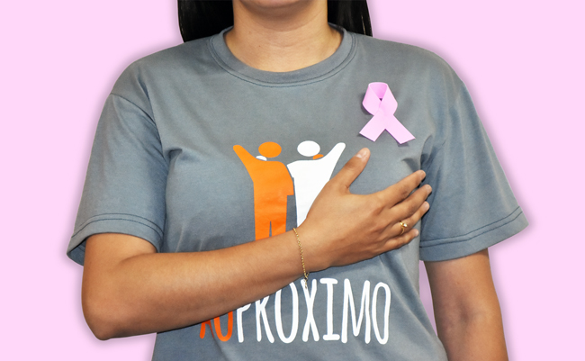 Mulher, em pé, vestido rosa, com a mão direita no seio esquerdo