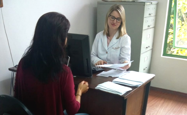 Em um consultório, a médica veste um jaleco branco está sentada atrás de uma mesa, com um computador a sua frente. A paciente está sentada do outro lado da mesa, vestindo vermelho 