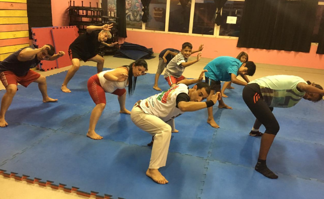 Em um tatame azul, grupo de pessoas jogando capoeira lado a lado