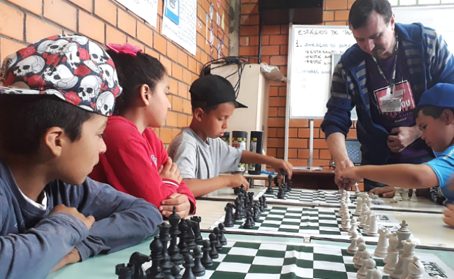 Em uma mesa, três meninos sentados com tabuleiros e peças de xadrez na suas frentes. Professo em pé ensino os movimentos.