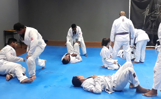 Em um tatame azul, jovens praticam  jiu-jitsu com quimonos brancos
