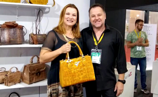 Prefeita em pé, segurando um bolsa, ao lado de um homem vestindo preto. Ao fundo, uma prateleira com bolsas