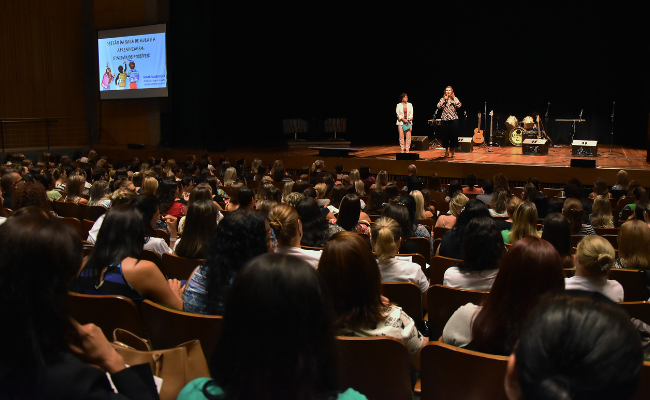 Ao fundo, um palco com a prefeita e a secretária em pé ao microfone. Auditório com pessoas sentadas
