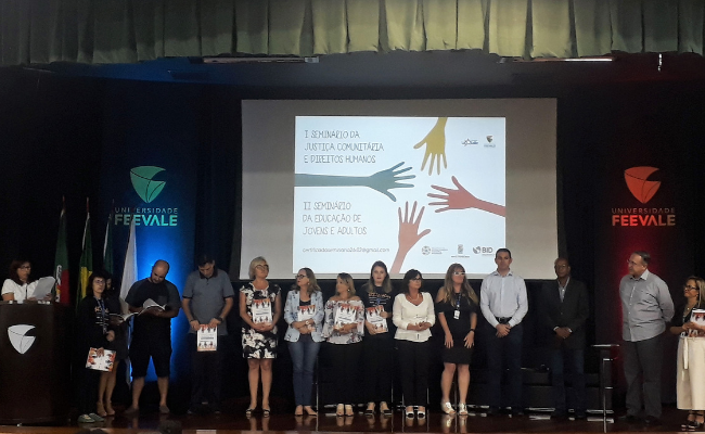 Em um palco, pessoas em pé com um telão projetado ao fundo