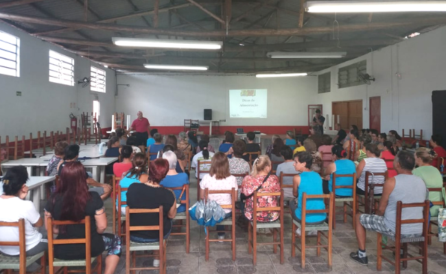 Ao fundo, Fagan em pé com as integrantes do PMI sentadas em cadeiras a sua frente