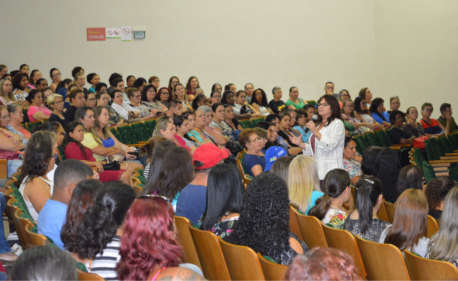 Em pé, secretária Maristela falando ao microfone e público sentado no entorno