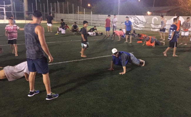 Em uma quadra de grama sintética, meninos fazem exercício espalhados