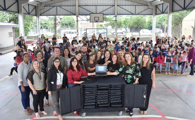 Com os chromebooks à frente, prefeita, secretária e grupo de pessoas posicionam-se em pé