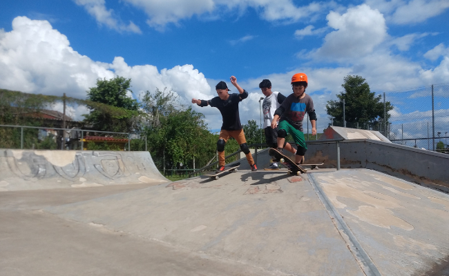 Em uma pista de skate, três meninos descem de uma rampa em cima do skate