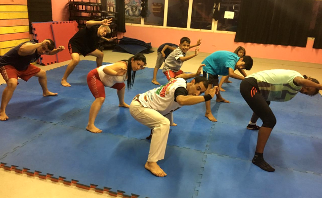 Em um tatame, jovens jogam capoeira um ao lado do outro