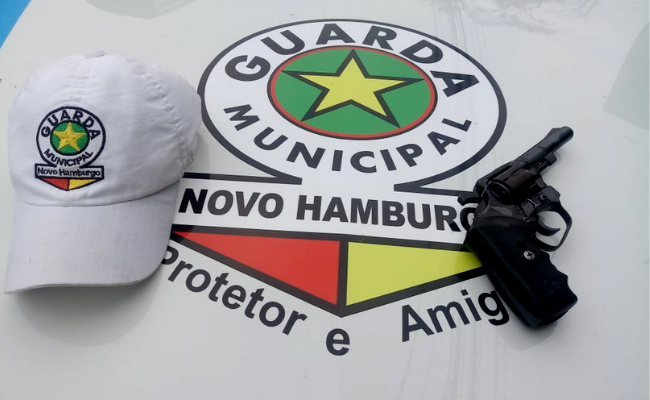 Arma preta em cima do capô de um veículo com a insígnia da Guarda Municipal desenhada. Ao lado, um boné também com a insígnia da Guarda Municipal