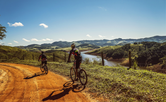 Em uma estrada de terra, dois homens andando de bicicleta. No horizonte, montanhas fecham o cenário