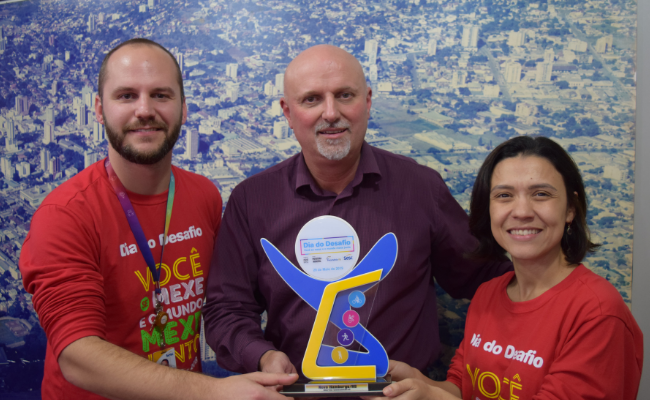 Três pessoas em pé, sendo dois homens e uma mulher à direita, segurando um troféu