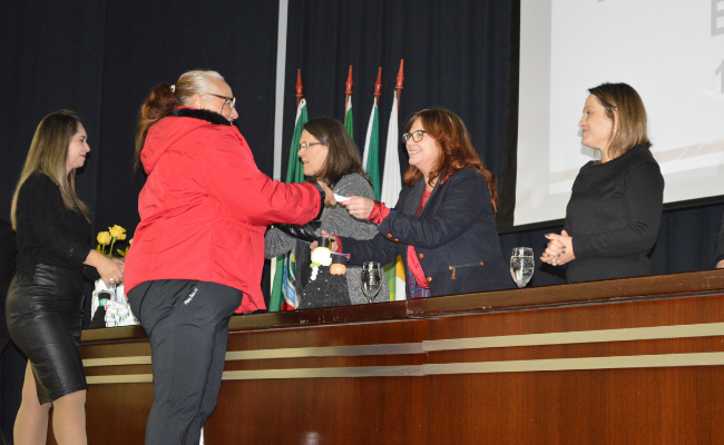 Em um palco, a secretária Maristela entrega um diploma para uma aluna