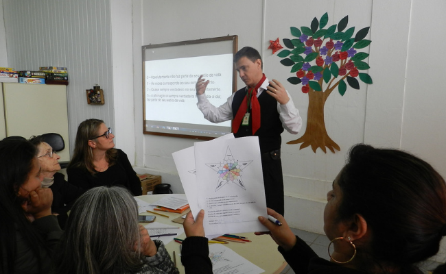 Em uma sala, homem em pé vestido com roupas da cultura gaúcha e quatro mulheres sentadas a sua frente, sendo que uma delas segura uma folha de papel com a imagem de uma estrela
