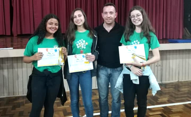 Três meninos vestindo uma camiseta verde e segurando um diploma, junto com o professor orientador