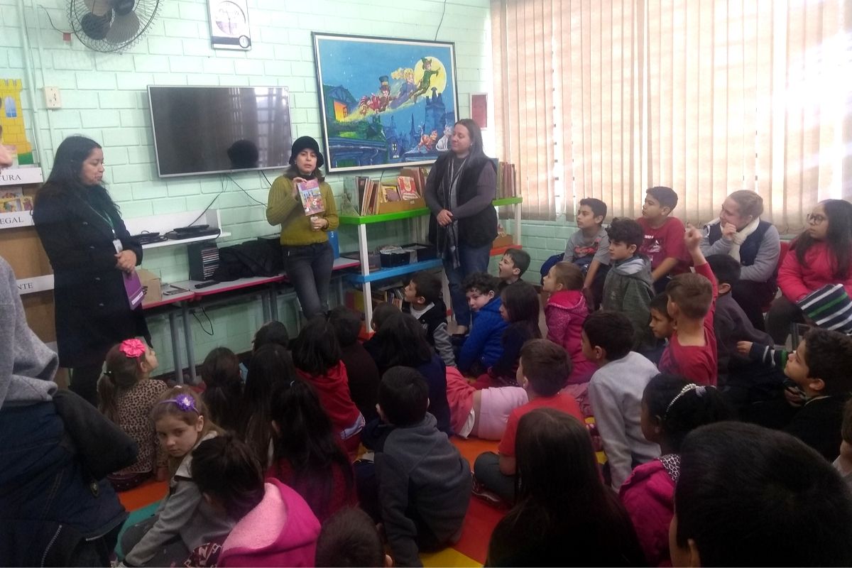 Em uma sala de aula, alunos sentados no chão e mulher em pé falando para estes alunos