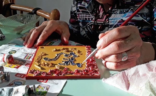 Mãos de mulher segurando um pincel e pintando em uma madeira quadrada pequena