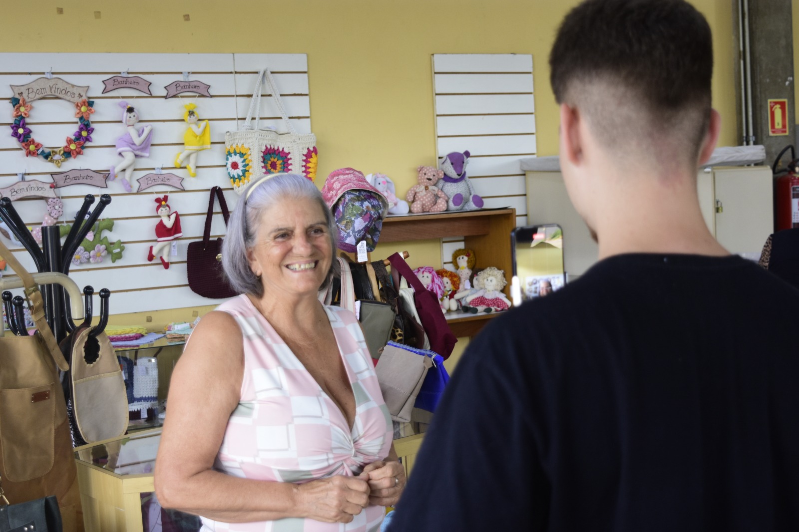 A imagem mostra um ambiente de loja que exibe produtos artesanais. Nas prateleiras e paredes estão expostos itens diversos, como bonecas de tecido, bolsas, chapéus e objetos decorativos.