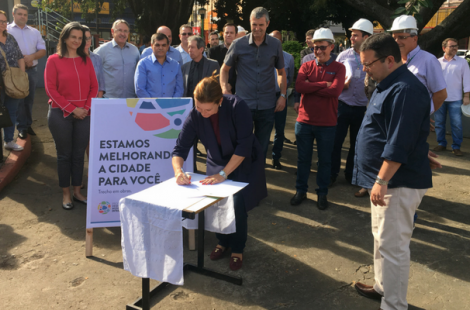Prefeita Fátima em pé assina o documento que está em uma mesa. Público ao redor assiste a assinatura 